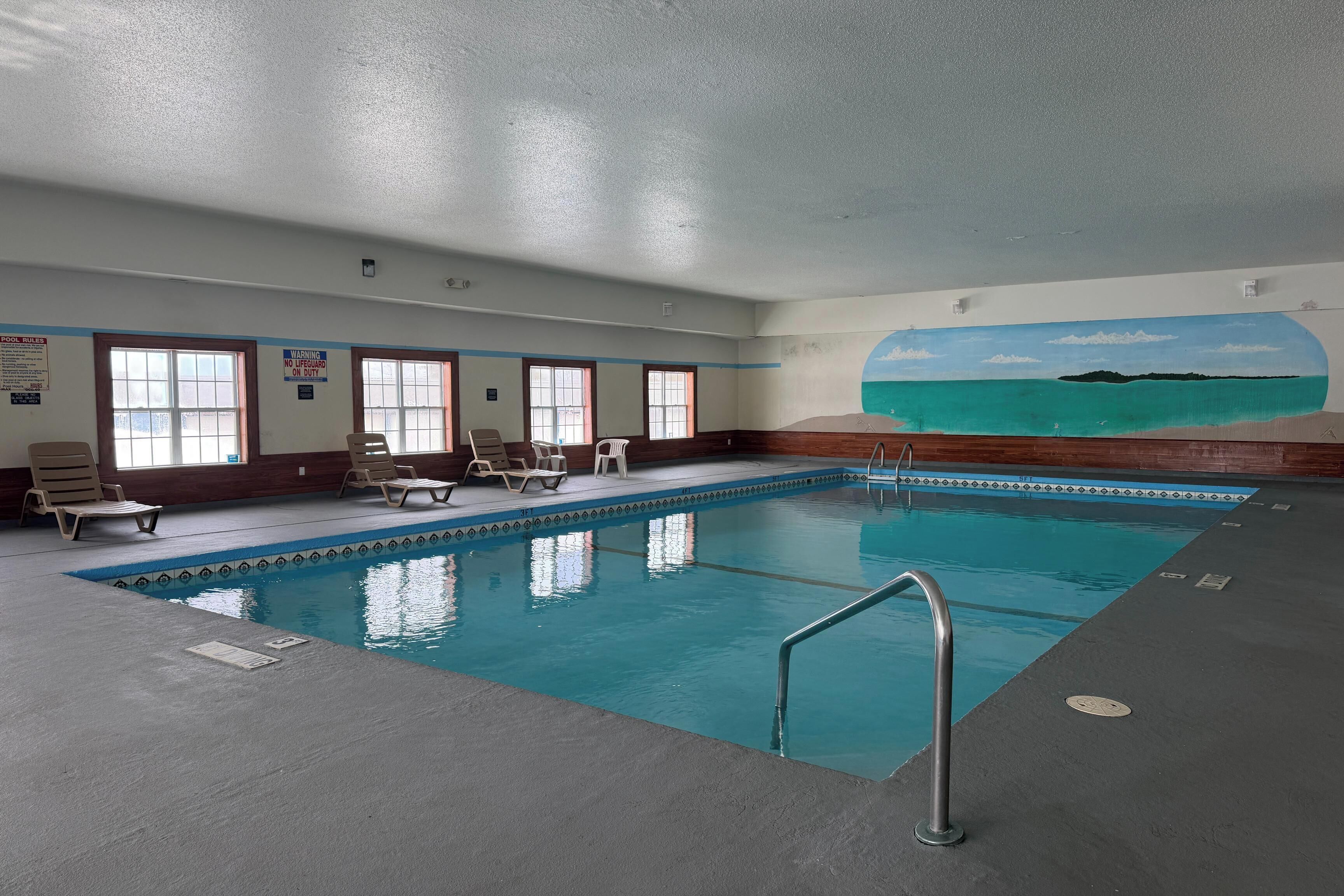 Una piscina interior con agua cristalina, rodeada de sillas de descanso y un mural con una escena de playa.