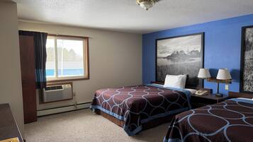 A hotel room with two beds, a large window, an air conditioner, a desk with a lamp, and a framed picture on the wall.