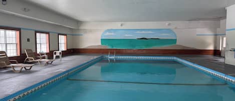 Una piscina interior con un mural de una playa, sillas de descanso y una terraza de madera.