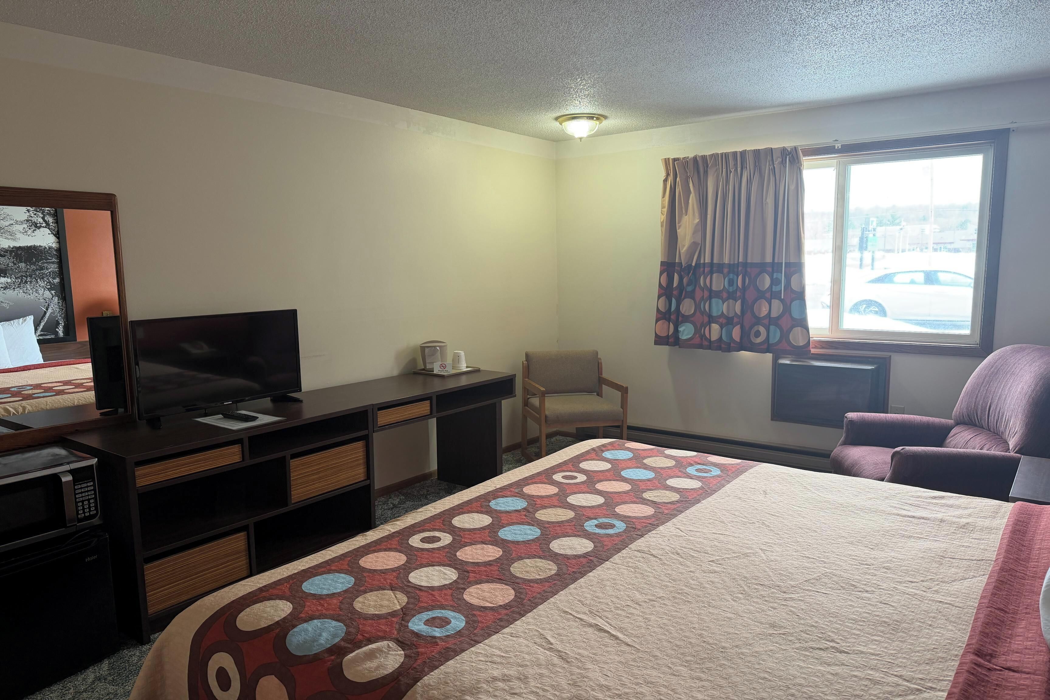 A hotel room with a bed, a desk with a TV, a chair, and a window with curtains.