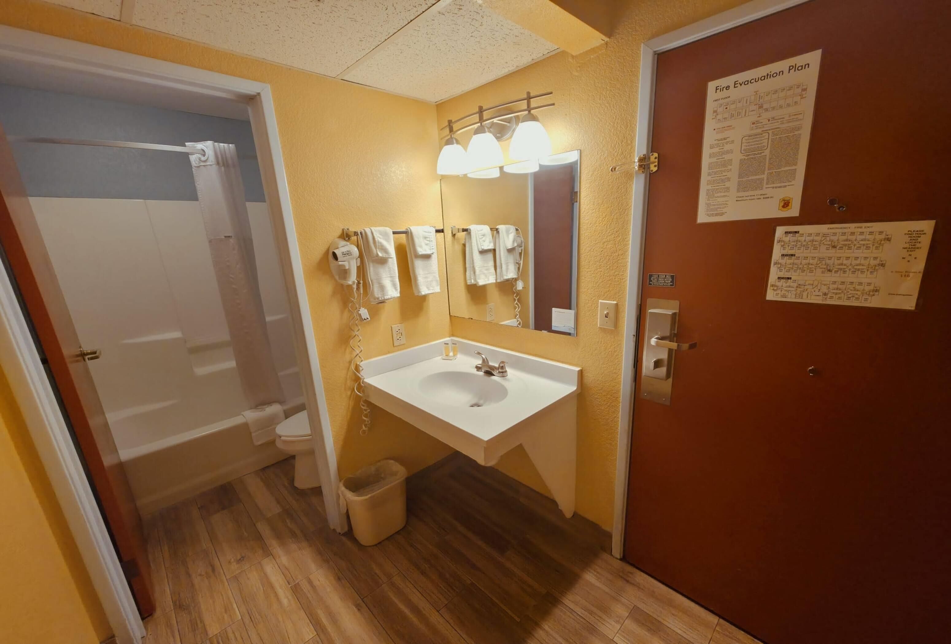 A bathroom with a white sink, mirror, and towel rack. There is a door with a fire evacuation plan on the wall.