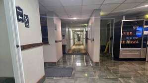 Ein Hotelgang mit einer Verkaufsmaschine, einem Mülleimer und Hinweisschildern für Aufzug und Treppe.