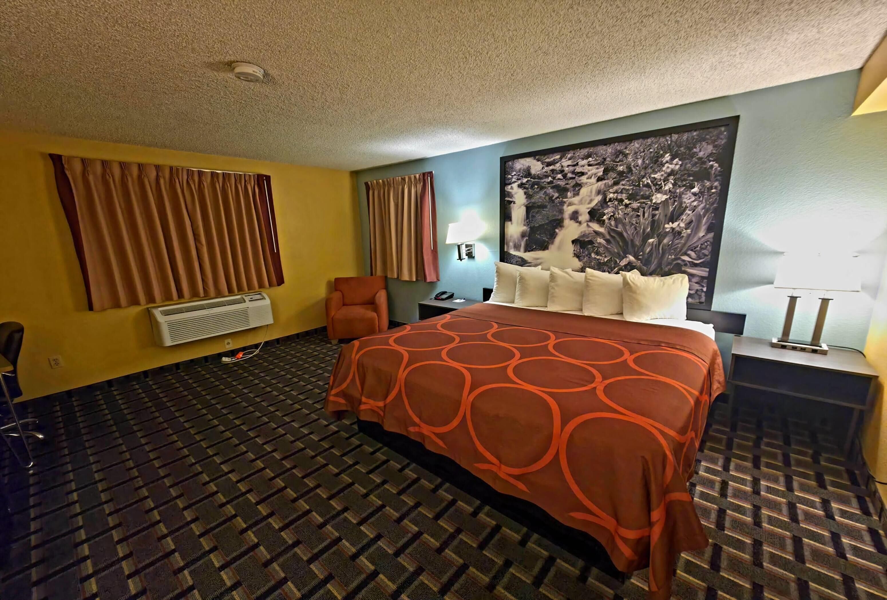 A hotel room with a patterned carpet, a bed with an orange circular design, a chair, a desk with a lamp, and a large wall painting.