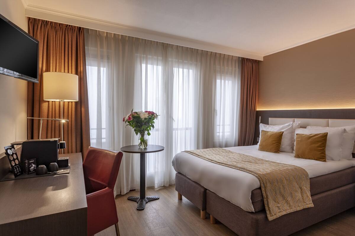A hotel room with a bed, a desk with a computer, a chair, a table with a vase of flowers, and a TV.