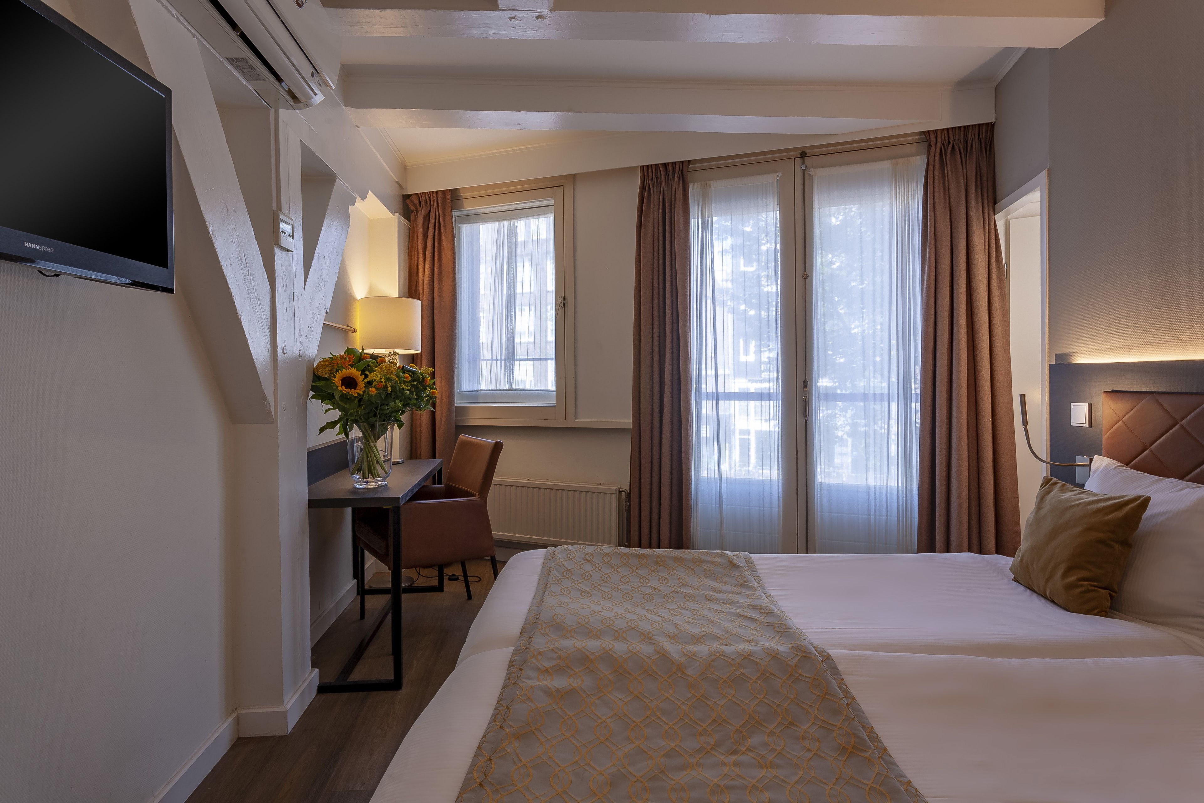 Une chambre d’hôtel avec un lit, un bureau, une chaise, une télévision et une fenêtre avec des rideaux.