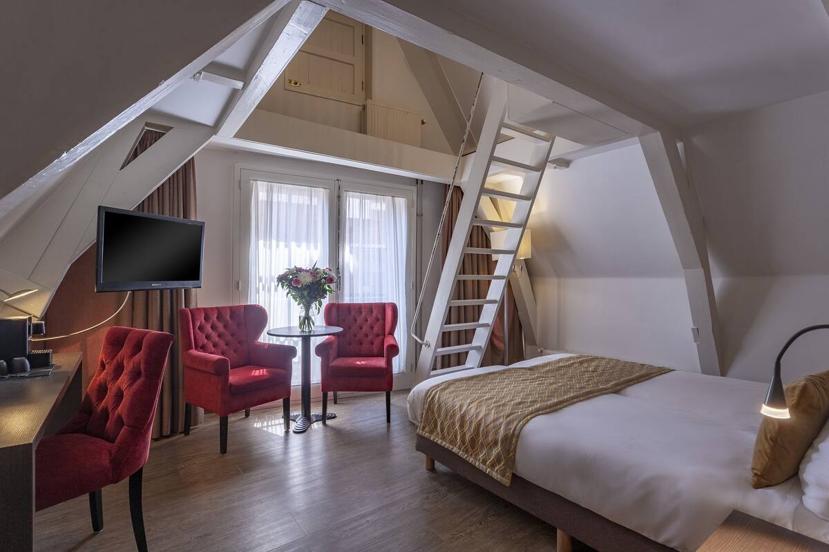 A bedroom with a bed, a desk, two red chairs, a small table with a vase, and a TV mounted on the wall.