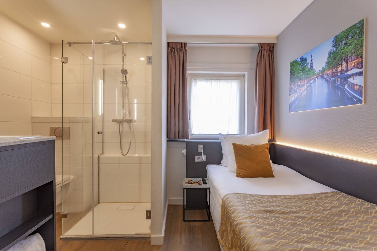 A hotel room with a glass-enclosed shower, a bed with a patterned bedspread, a nightstand with a book, and a window with curtains.