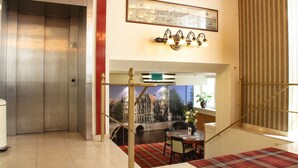 A hotel lobby with an elevator, a staircase, a dining area with a table and chairs, and a cityscape mural on the wall. - Hotel Avenue (Amsterdam)