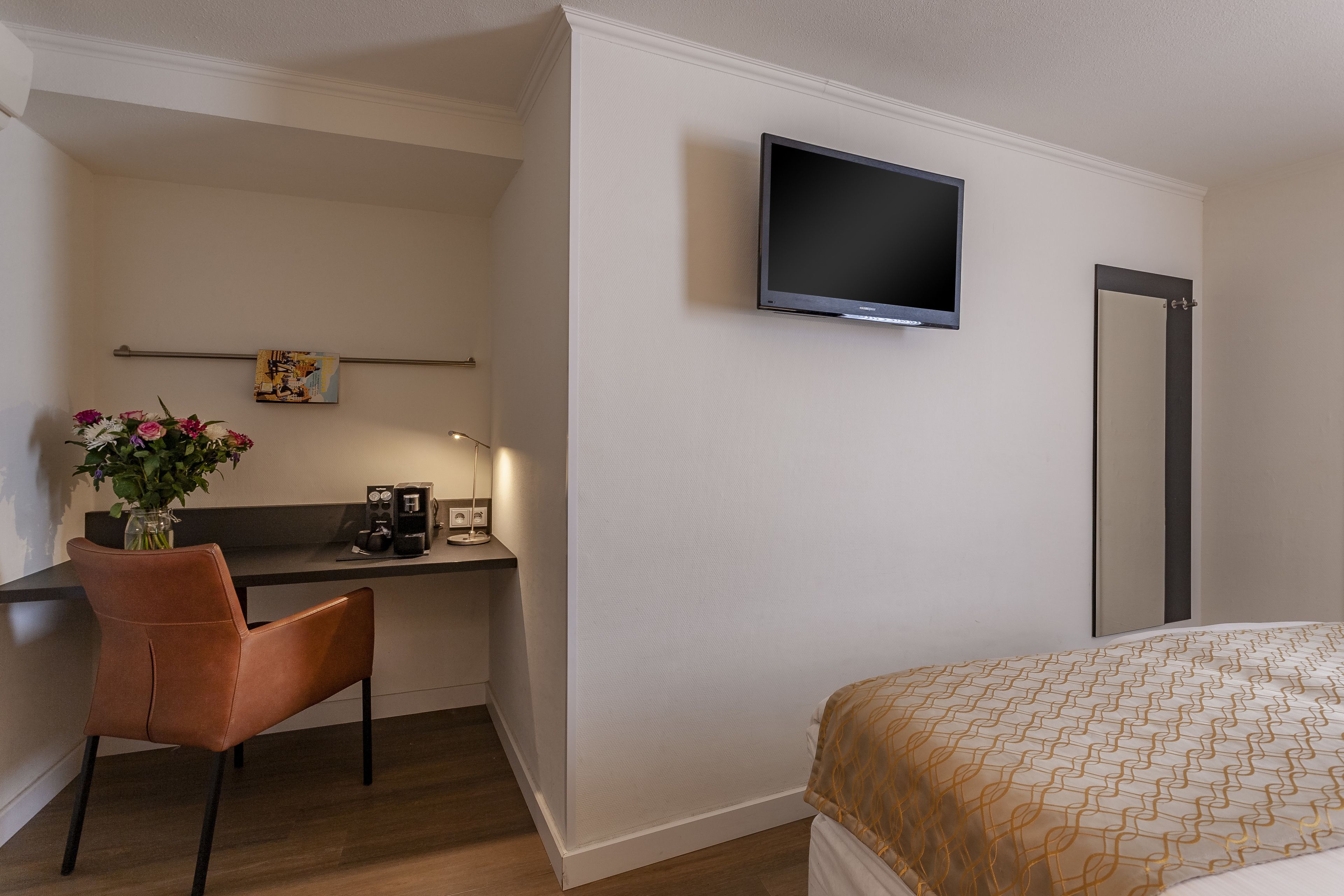 A hotel room with a bed, a desk with a chair, a television mounted on the wall, and a small artwork above the desk.