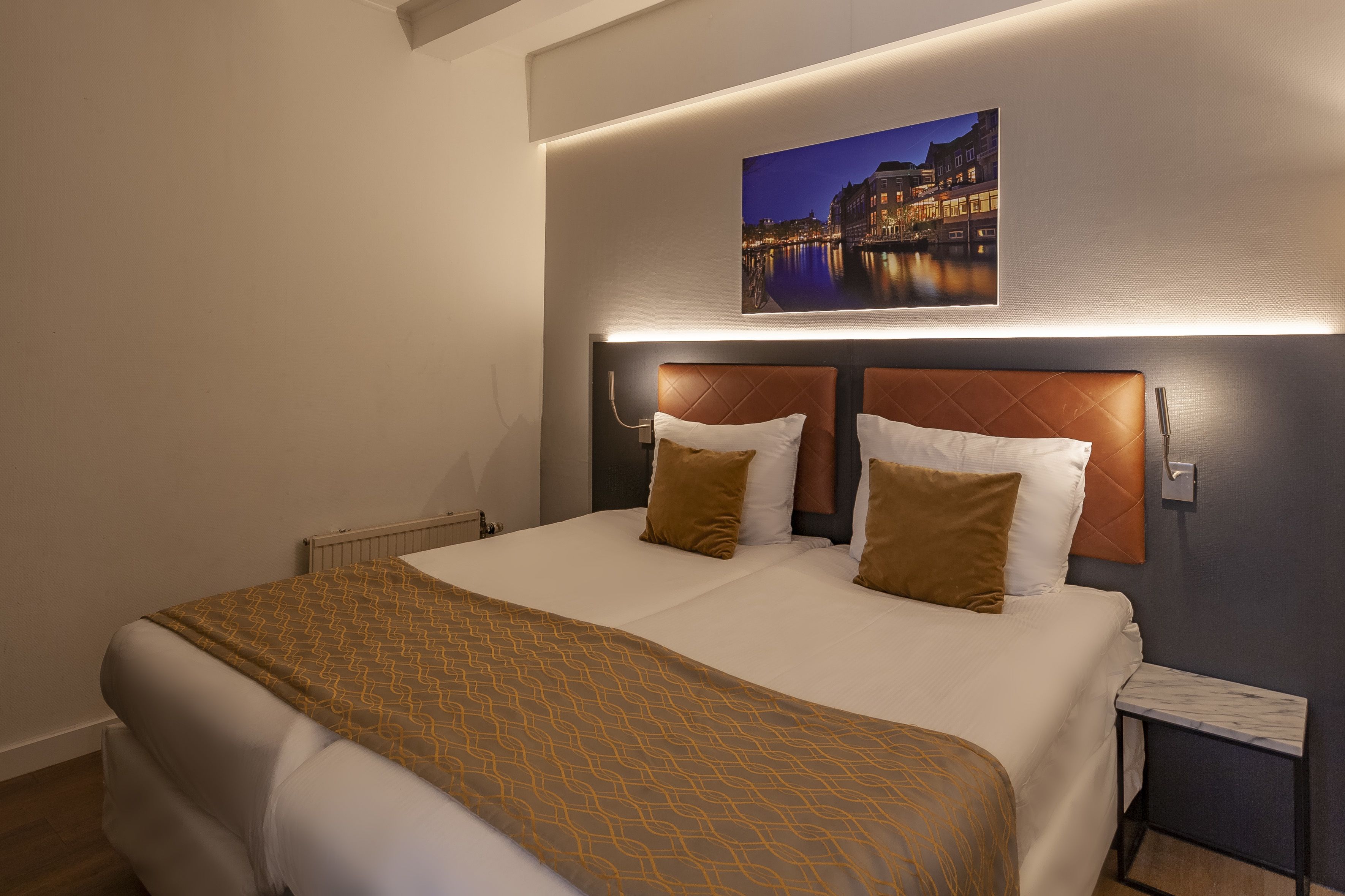Een hotelkamer met een groot bed, een hoofdeinde met een ingebouwde nachtkastje en een aan de muur hangend stadsbeeld.