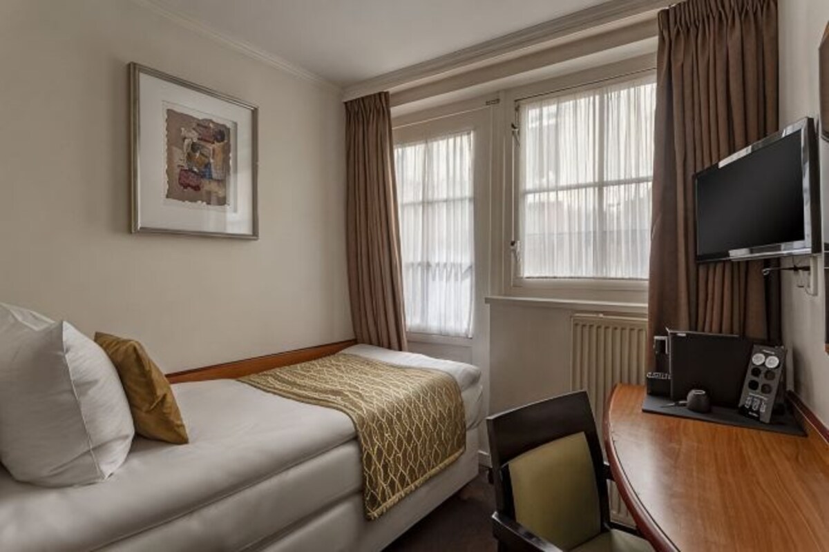 A hotel room with a bed, a desk with a computer, a TV, and a window with curtains.