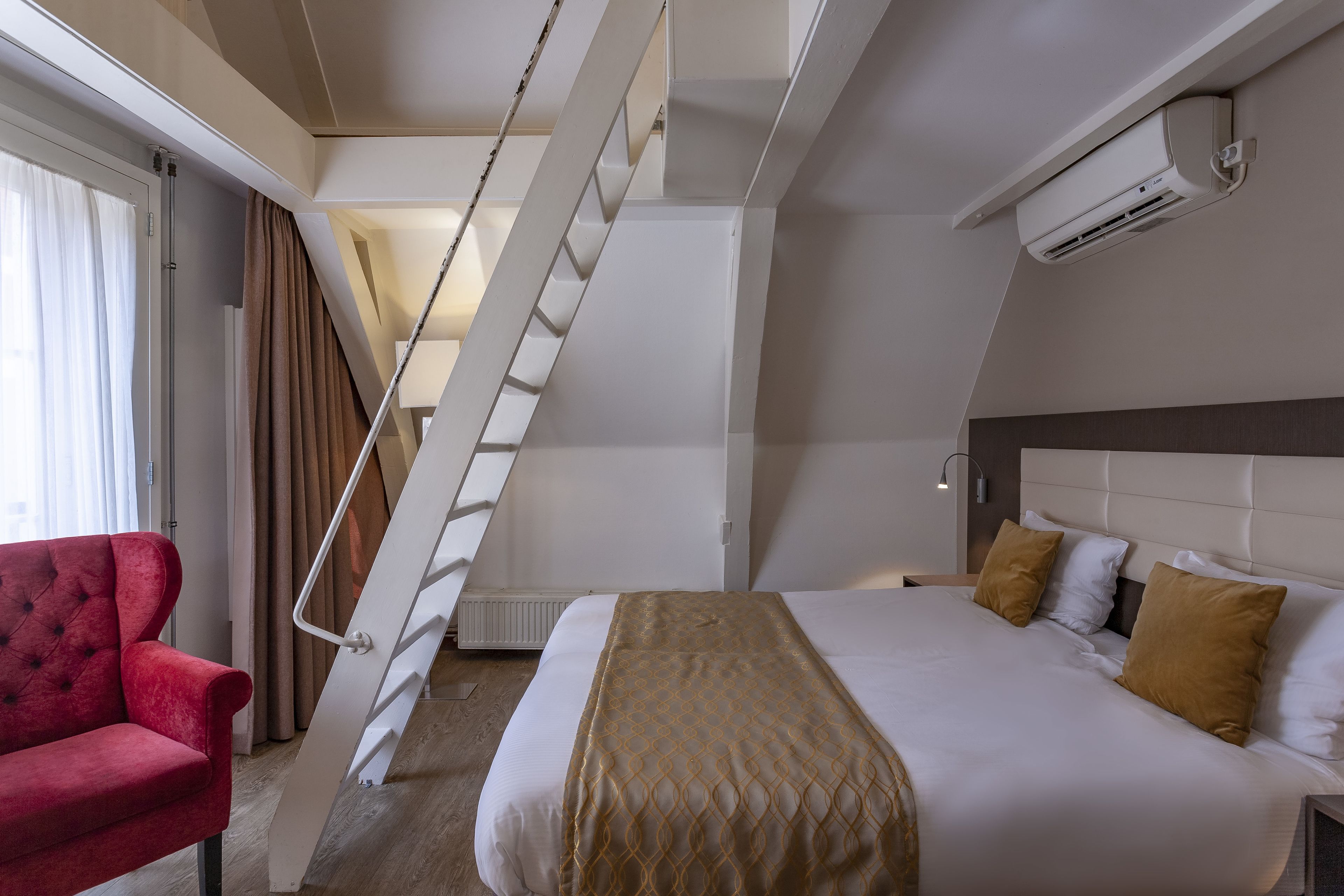 Una habitación de hotel con una cama, un sillón rojo, una escalera y un equipo de aire acondicionado.