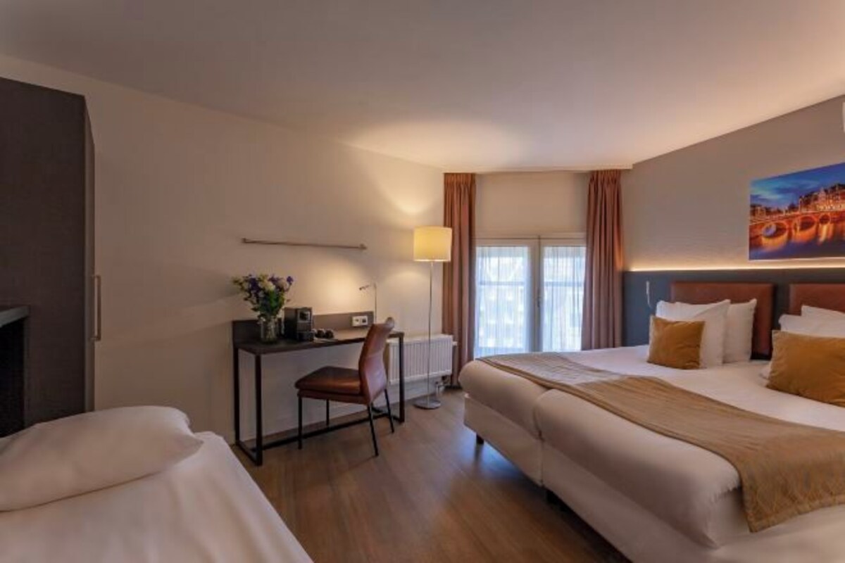 A hotel room with a large bed, a desk, and a chair. There is a window with curtains and a painting on the wall.