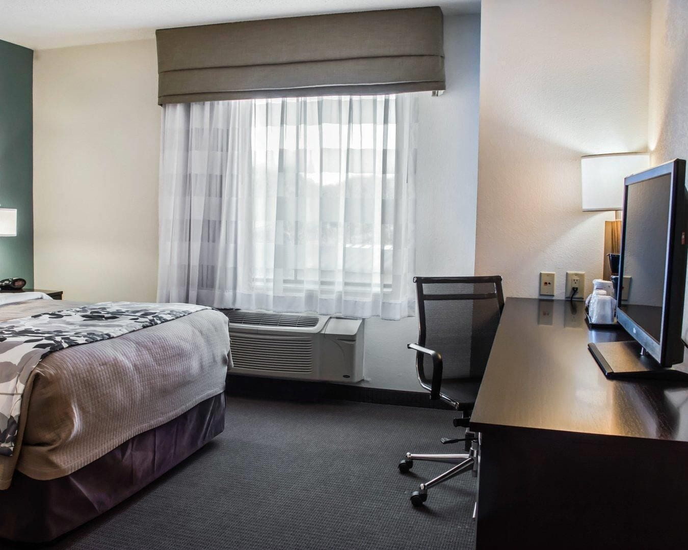 A hotel room with a bed, a desk with a computer, a chair, and a window with sheer curtains.