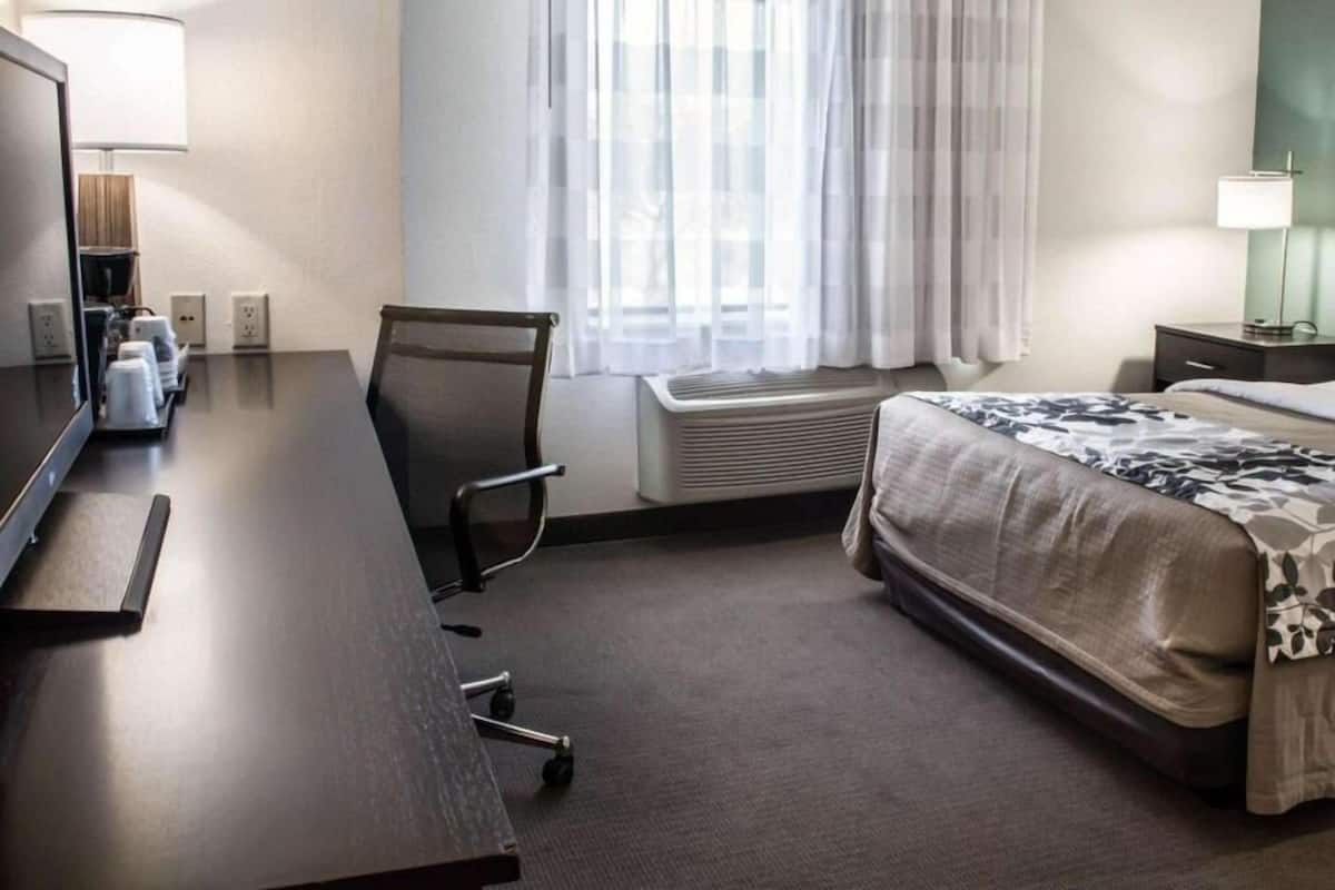 Une chambre d’hôtel comprenant un lit, un bureau avec une chaise, une fenêtre avec des rideaux, un appareil de climatisation et une lampe sur la table de chevet.