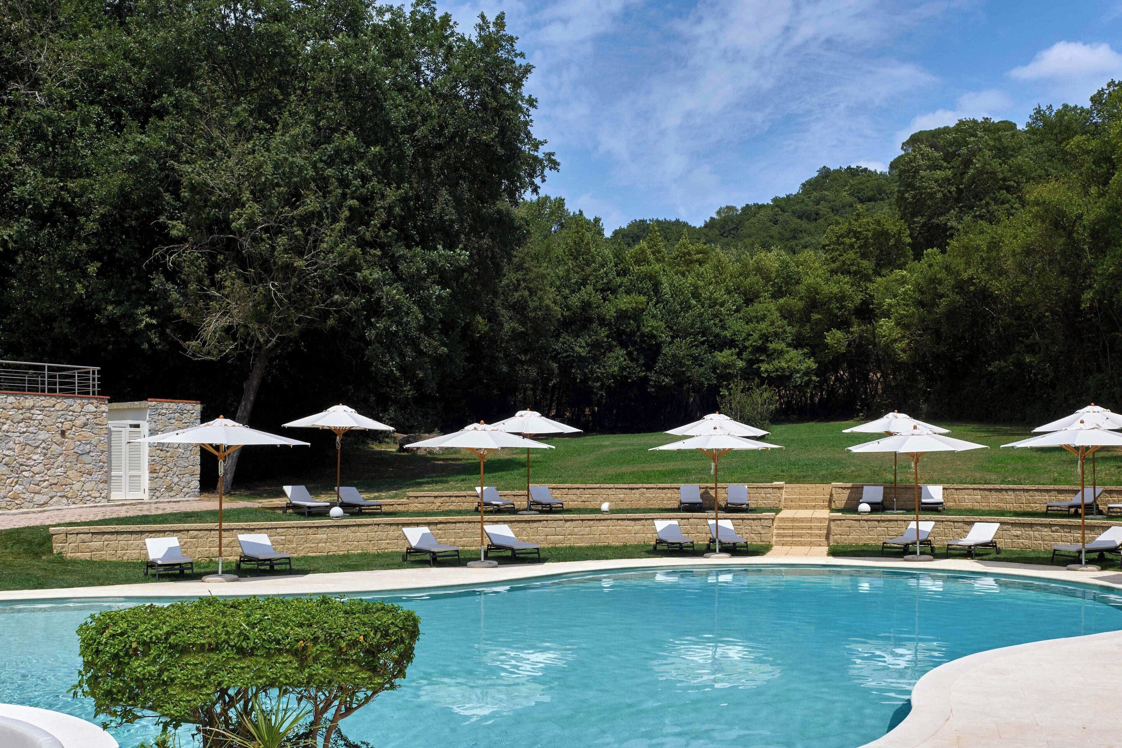 Une piscine entourée d’un mur de pierre et d’une végétation luxuriante, avec des chaises longues et des parasols à proximité.