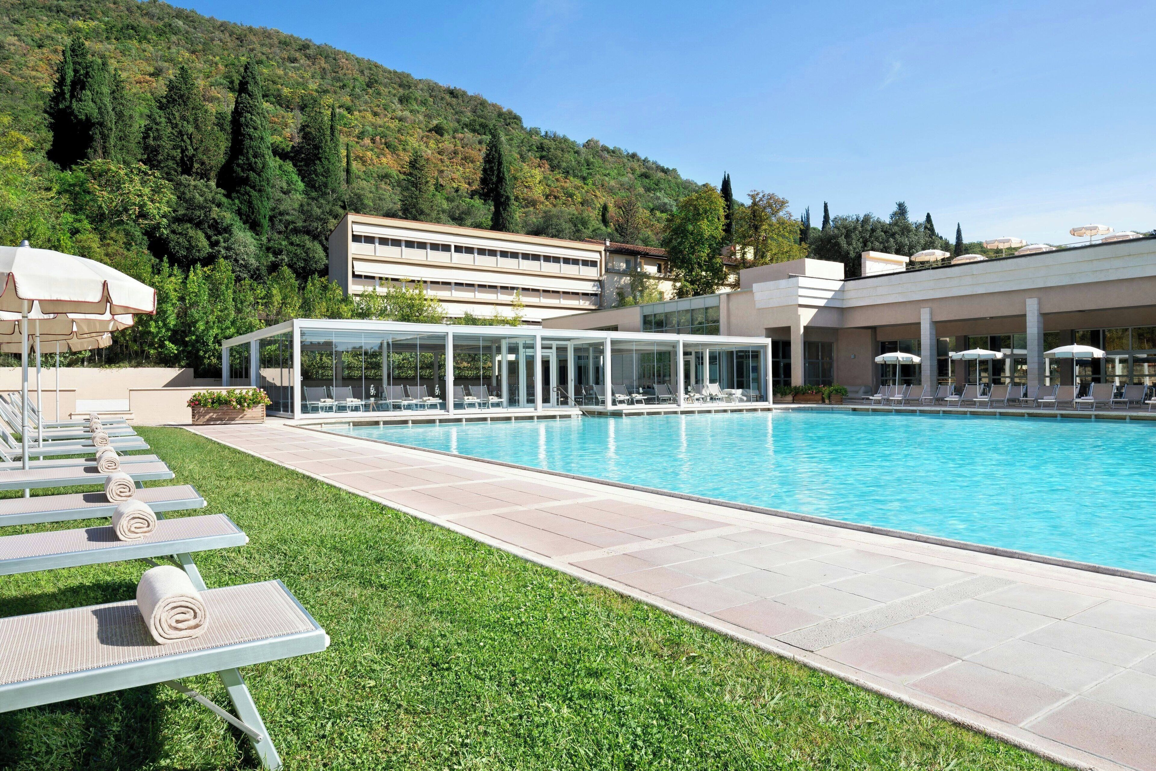 Una piscina con sillas de descanso y sombrillas, rodeada por un césped cuidado y un edificio de diseño moderno.