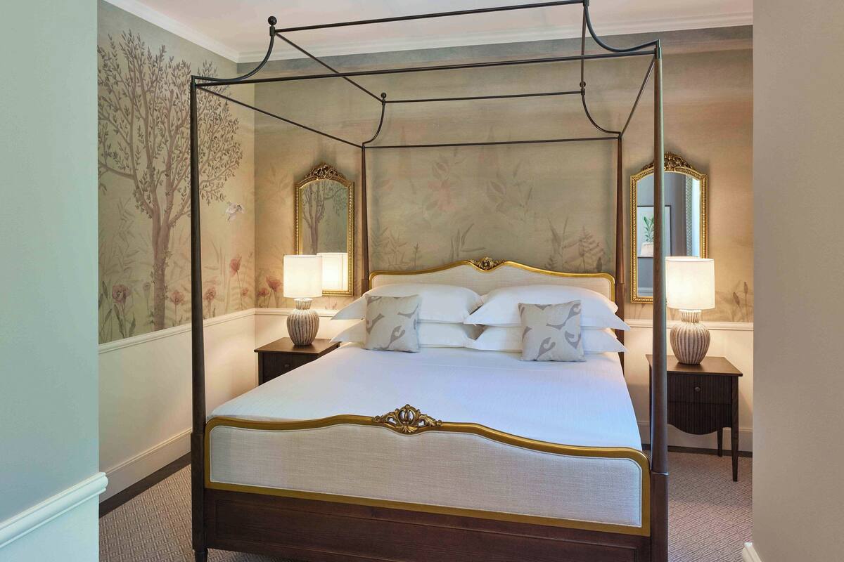 A four-poster bed with white linens and gold trim, flanked by two bedside tables with lamps, in a room with floral wallpaper and a large mirror on the wall.