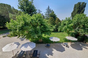 Een terras buiten met tafels en parasols, omgeven door bomen en een gazon.