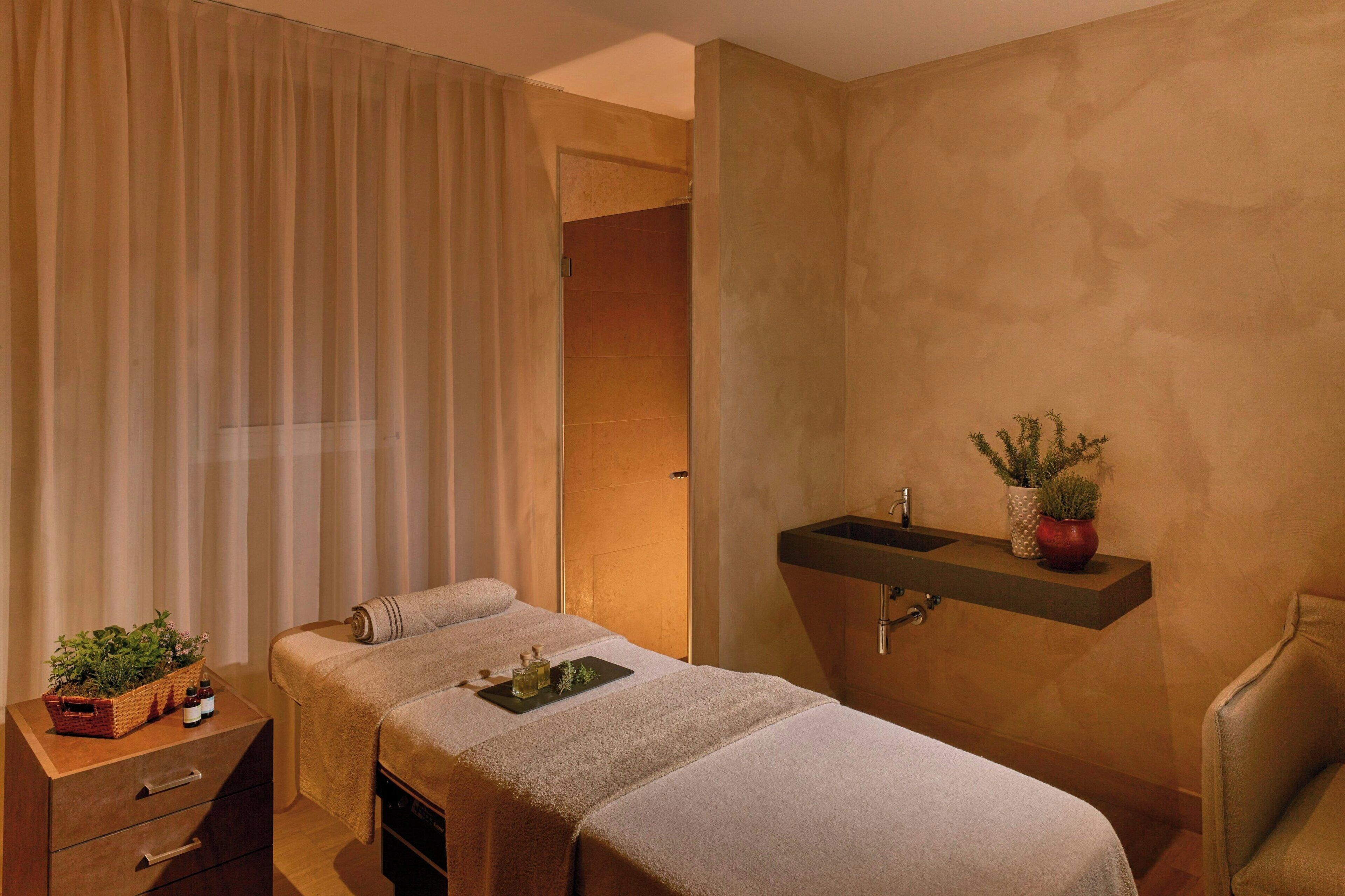 A hotel room with a bed, a small bedside table with a plant, a shelf with toiletries, and a shower area.