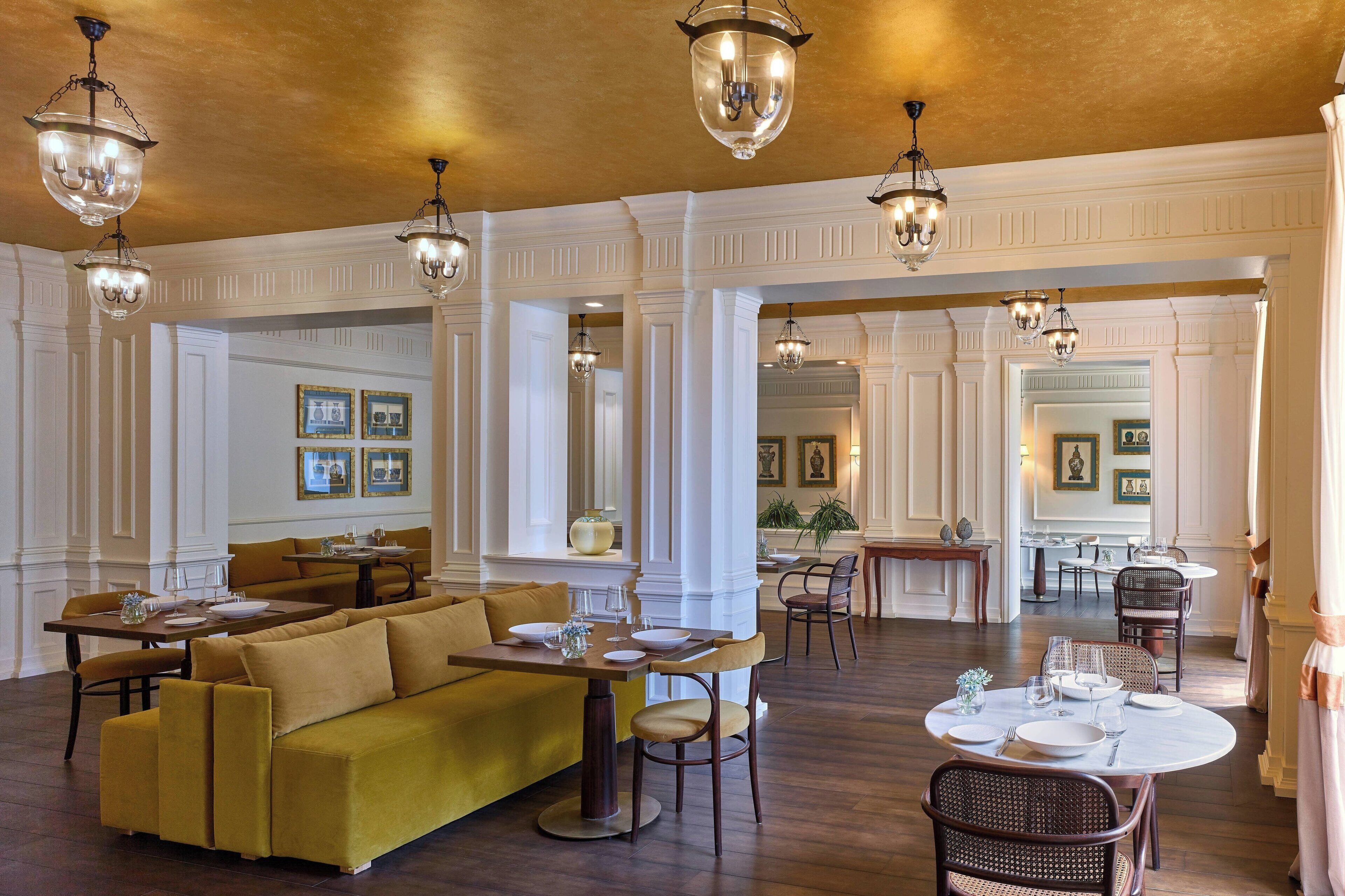 A spacious dining area with high ceilings, chandeliers, and a yellow sofa. Tables are set with plates and glasses.