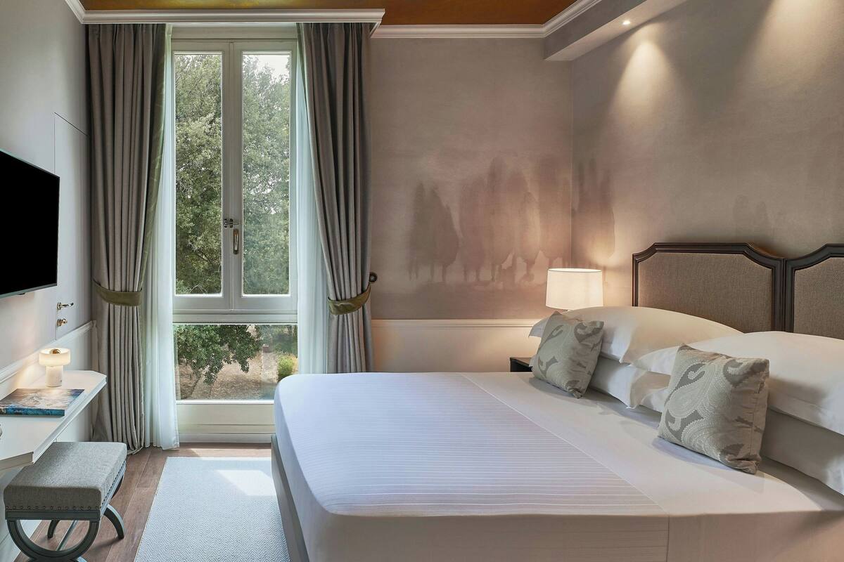 A hotel room with a large bed, a bedside table with a lamp, a TV mounted on the wall, and a window with curtains.