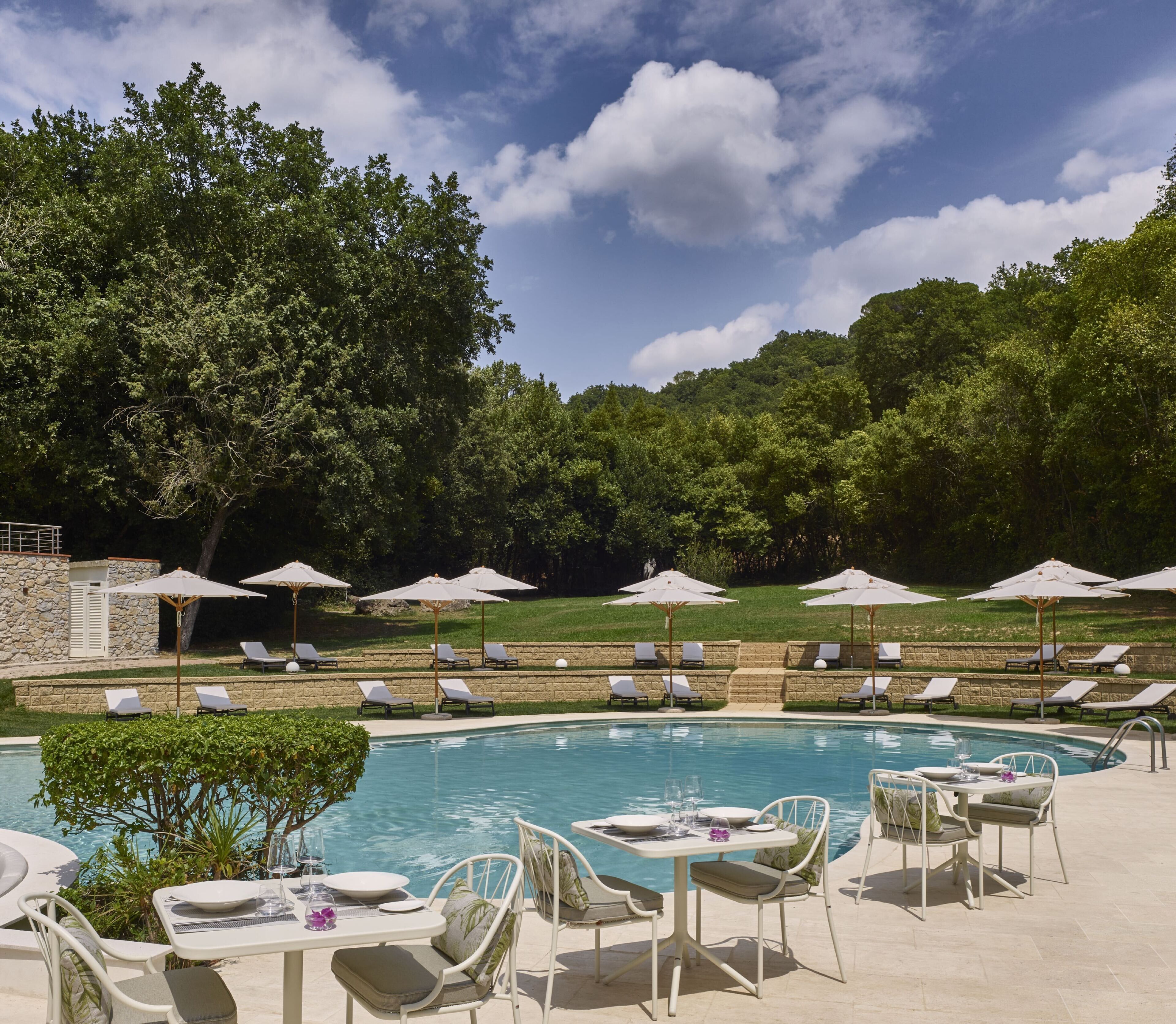 A pool area with lounge chairs and umbrellas, surrounded by trees and a stone wall.