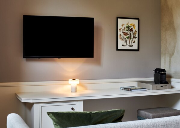 Une chambre moderne dotée d’un téléviseur à écran plat fixé au mur, d’une table de chevet avec une lampe, d’une illustration botanique encadrée et d’une machine à café.