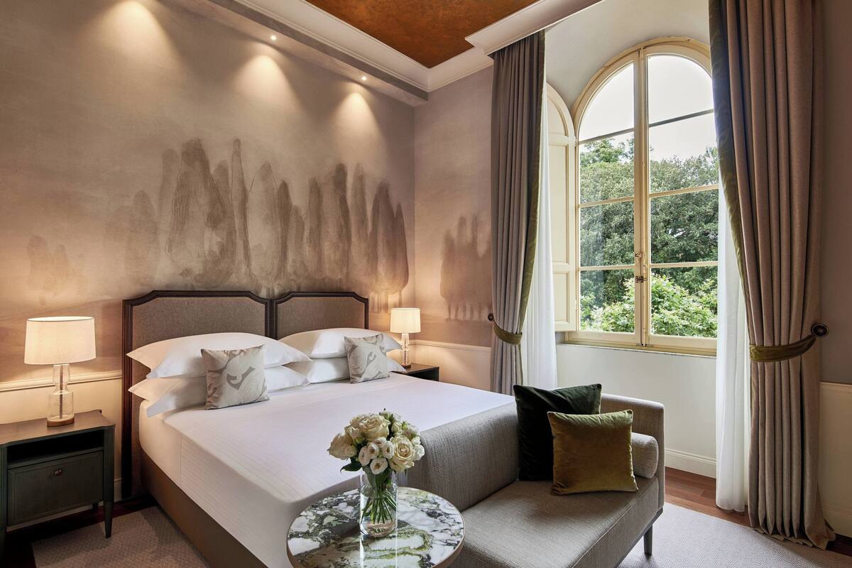 A hotel room with a large bed, a round side table with a vase of flowers, a sofa, and a window with curtains.