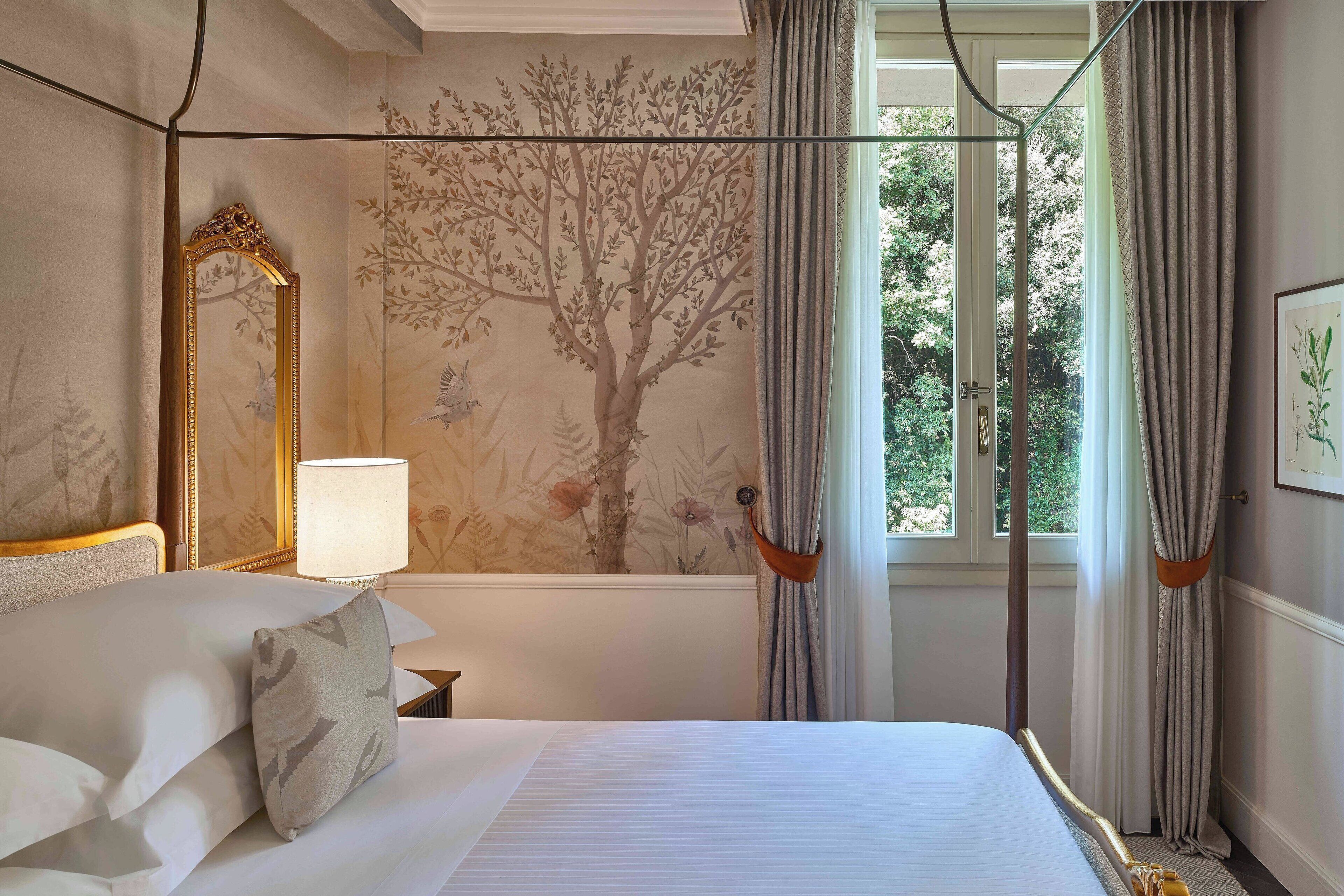 A bedroom with a canopy bed, a large mirror, a wall mural, and a window with curtains.