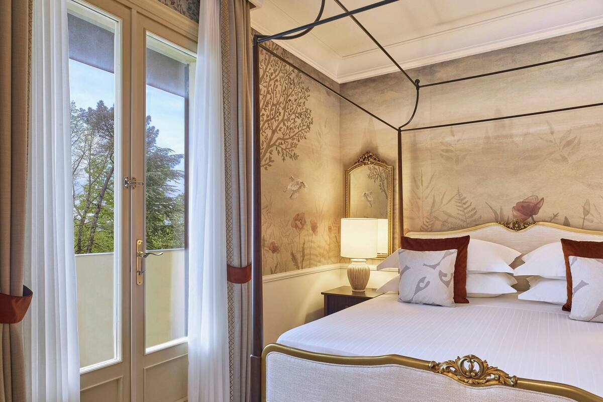 A hotel room with a large bed, a nightstand with a lamp, and a view of trees through the window.
