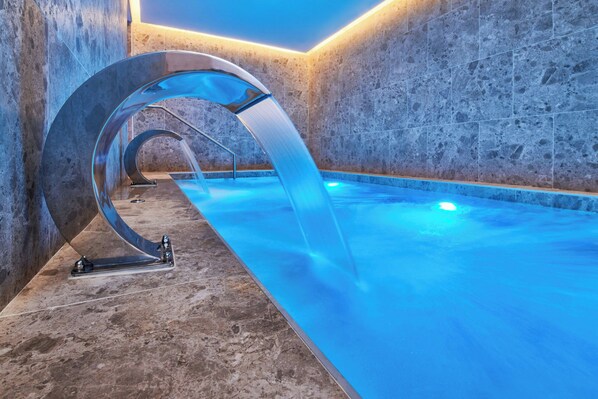 Una piscina interior moderna con una original fuente curva y una pared revestida de piedra.