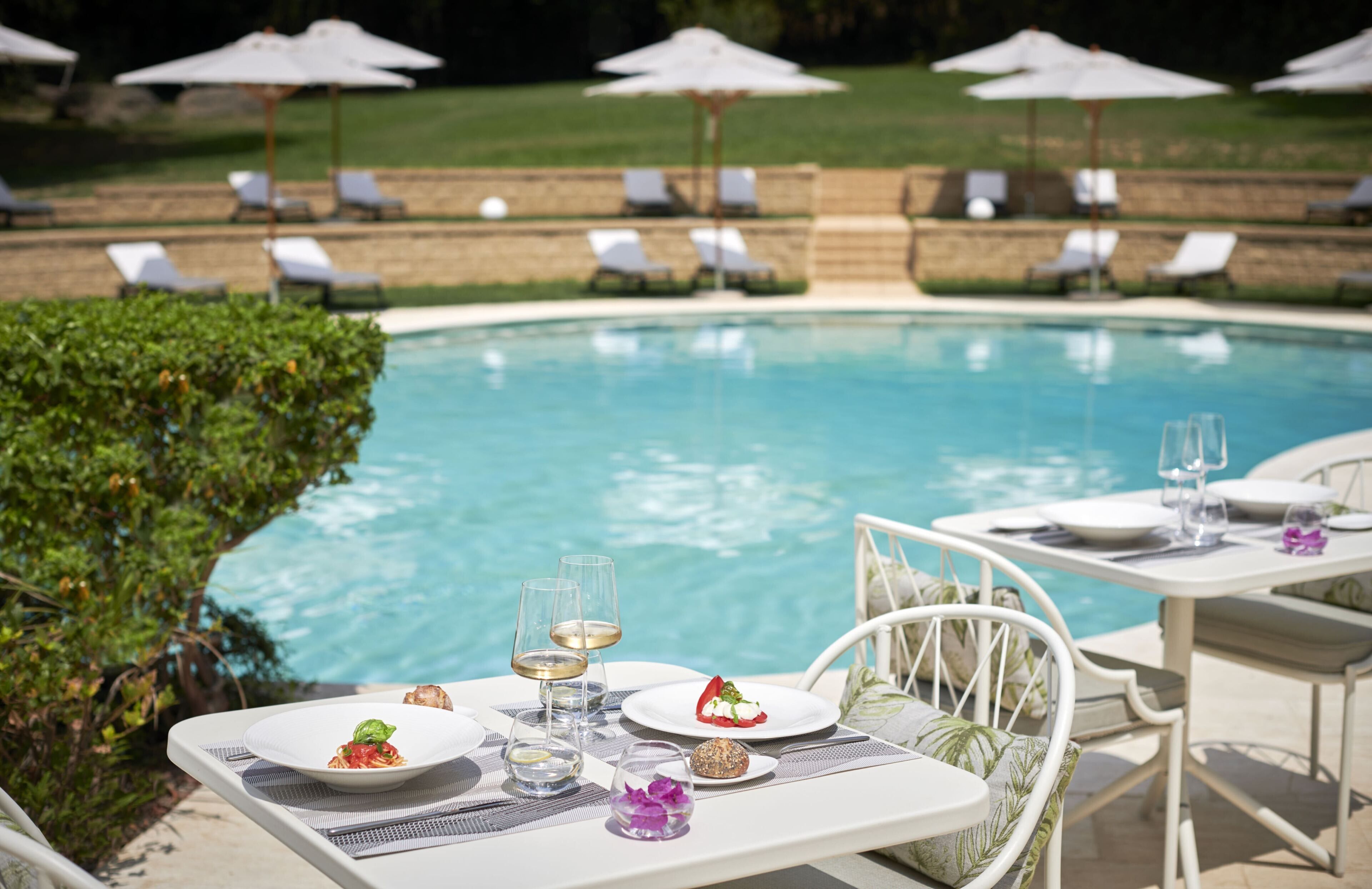 A dining table set with plates, glasses, and a dessert, overlooking a pool with lounge chairs and umbrellas.
