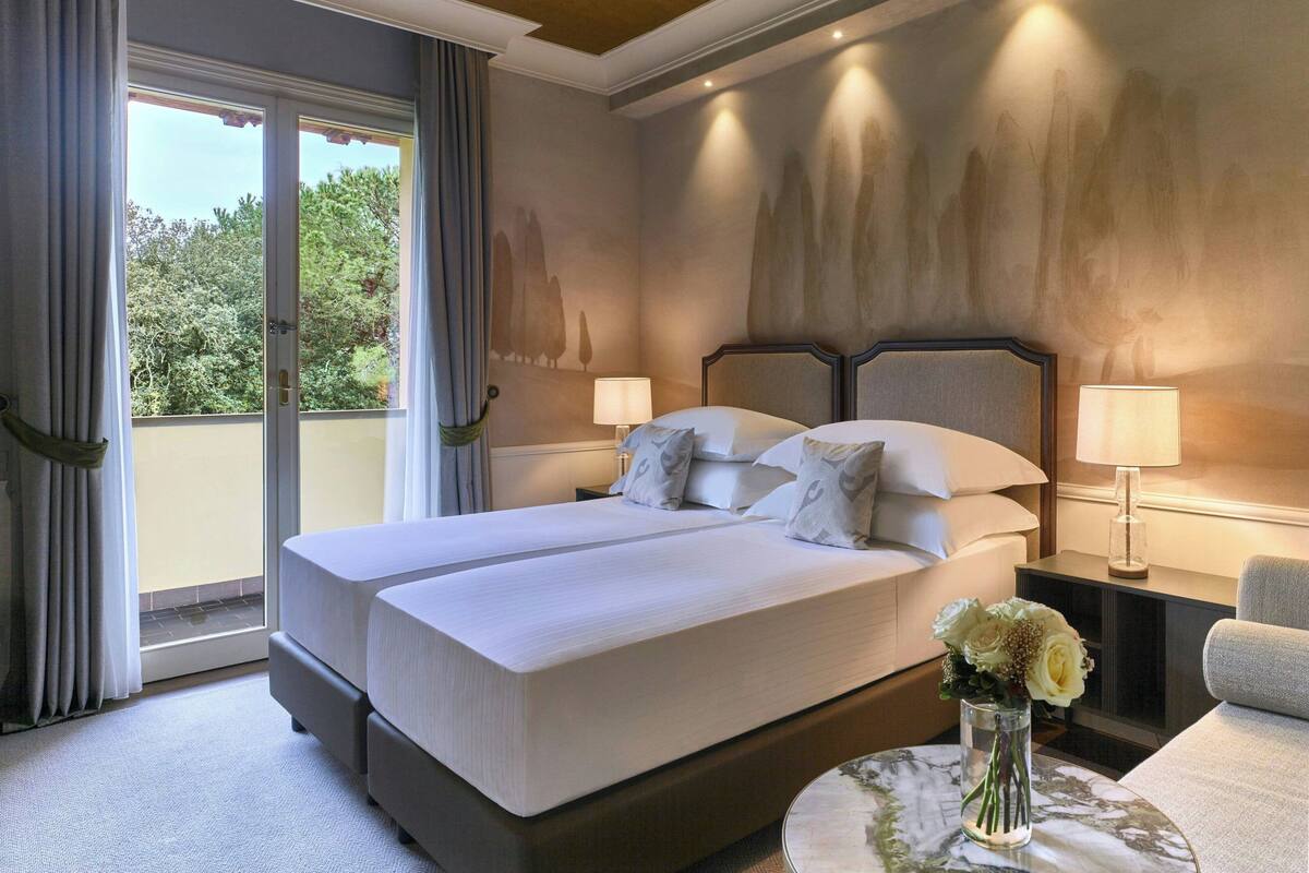 A double bed with a headboard, two bedside tables with lamps, a vase with flowers, and a view of trees through a sliding door.