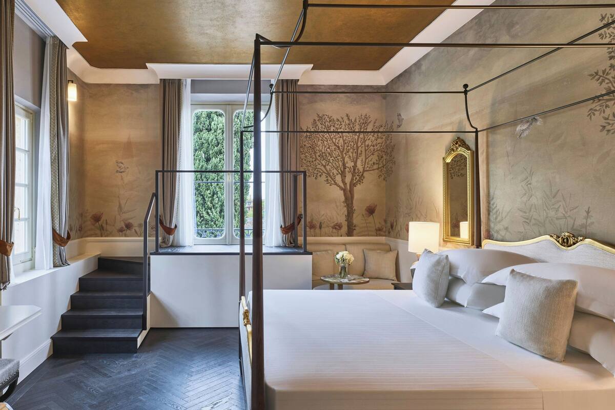 A bedroom with a large bed, a window with curtains, a staircase, and a wall-mounted mirror.