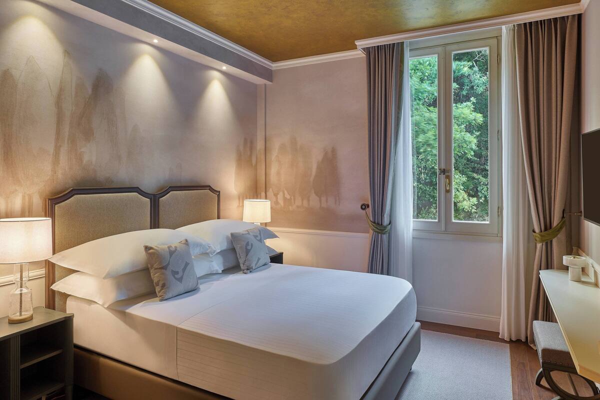 A neatly made bed with pillows, a bedside table with a lamp, a window with curtains, and a desk with a chair.