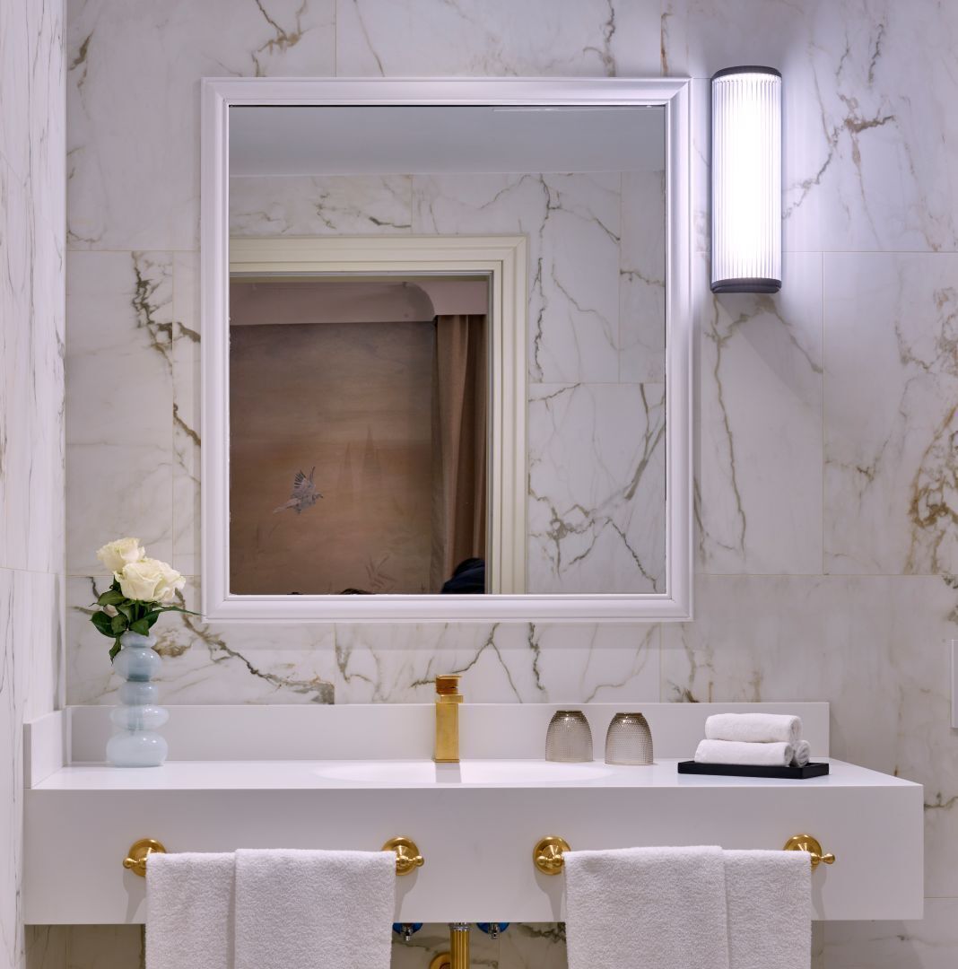 A bathroom with a white marble backsplash, a large mirror, a gold faucet, and two towel racks with white towels.