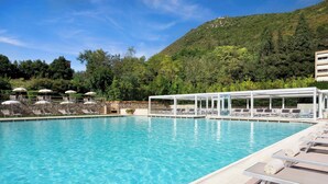 Une piscine d’un bleu éclatant, bordée de chaises longues et d’ombrelles, entourée de verdure et adossée à une colline.