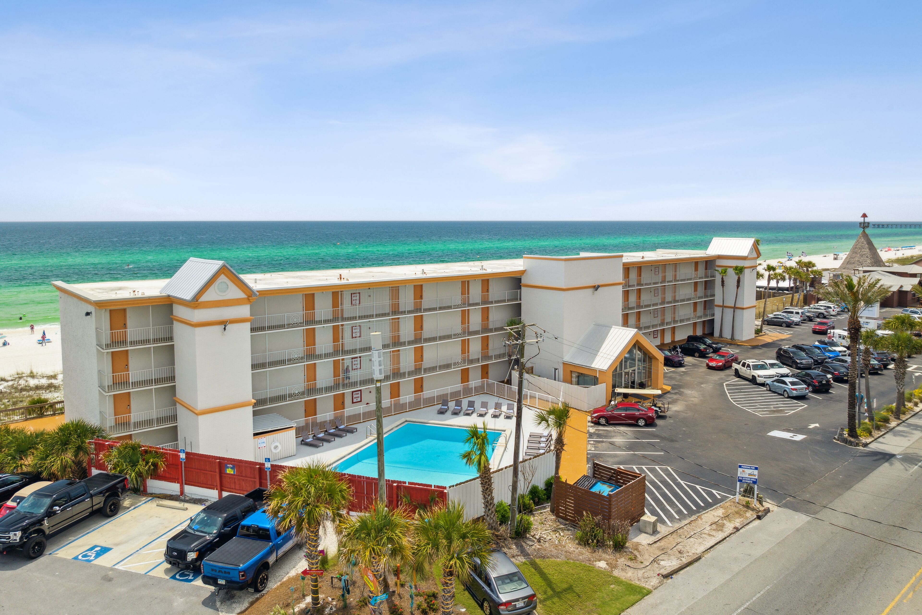 A hotel with a swimming pool, parking lot, and beach view.