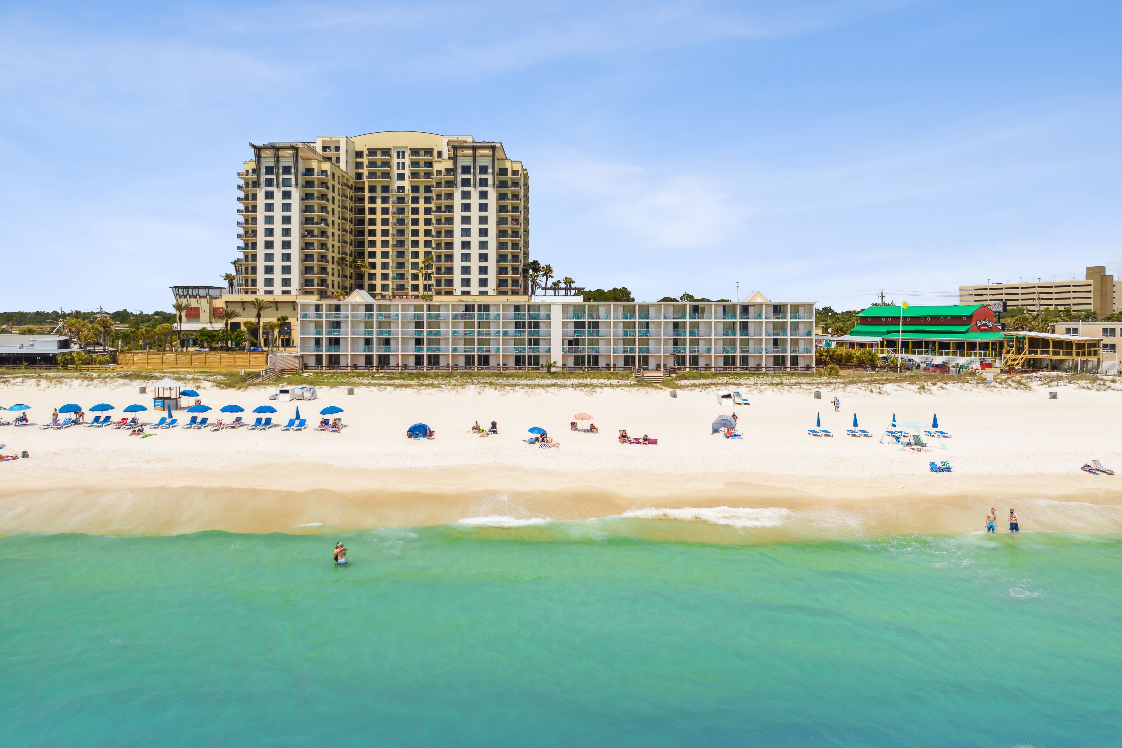 Un hotel frente a la playa con un edificio grande, una playa de arena y varias sombrillas.