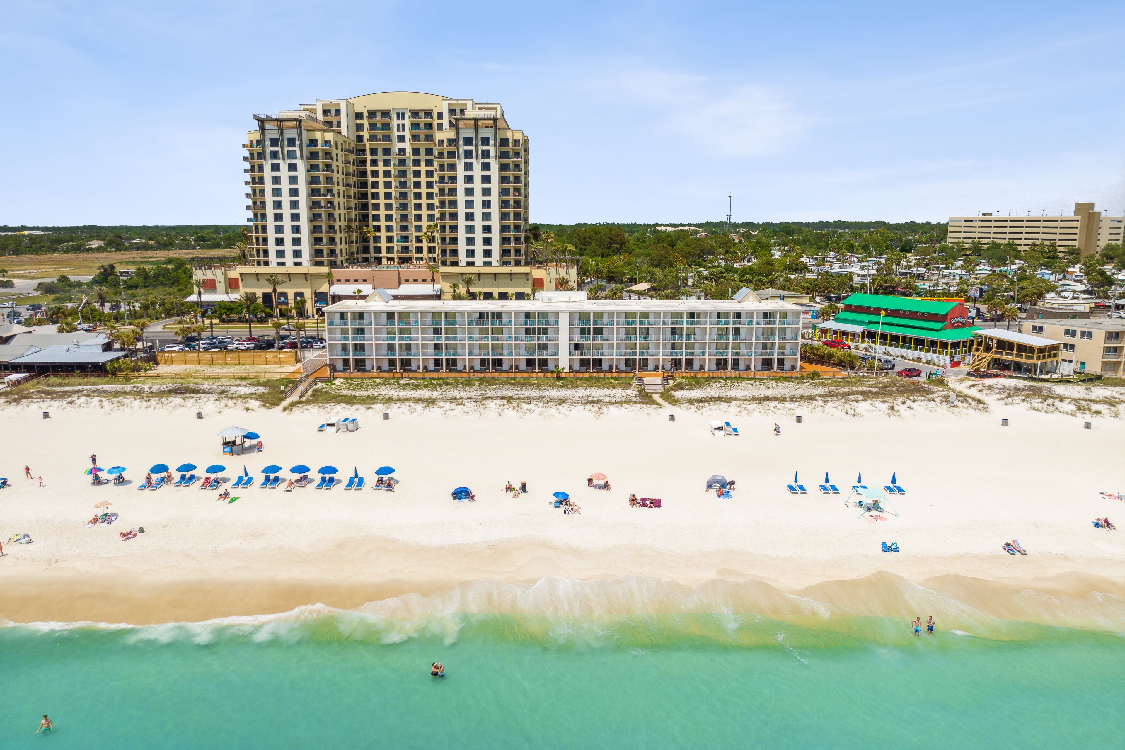 A beachfront hotel with multiple floors, a beach area with umbrellas and lounge chairs, and a view of the ocean.