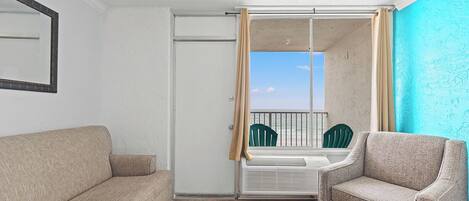 A room with a beige sofa, a green chair, a sliding glass door leading to a balcony with a view, and a white radiator.