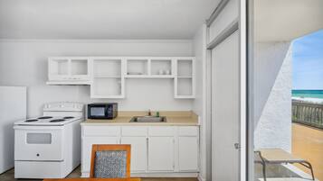 A compact kitchen with white cabinets, a stove, a microwave, and a sink. There's a balcony with a view of the beach.