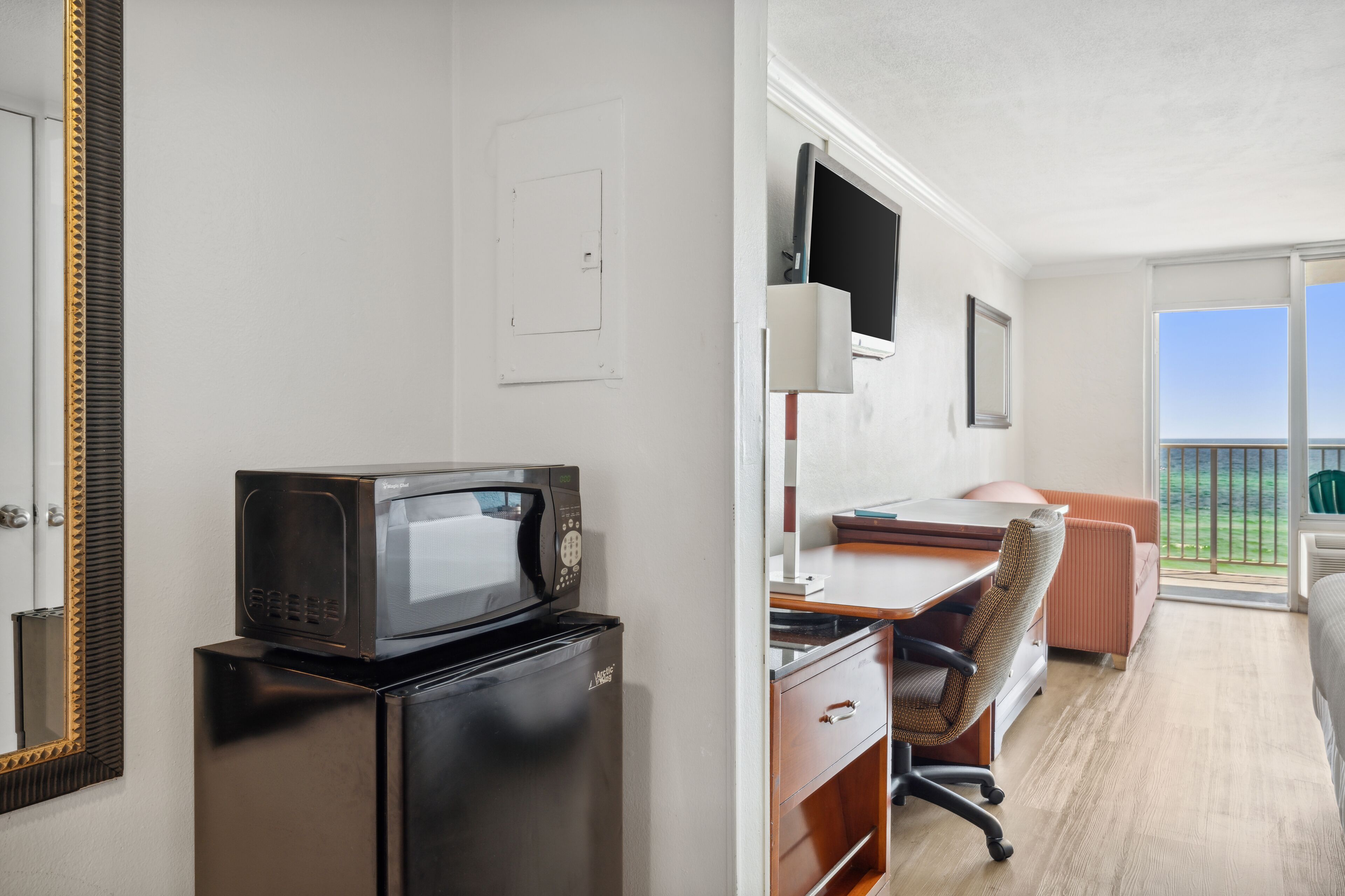 A compact hotel room with a kitchenette, a desk, a chair, a bed, and a large window with a view.