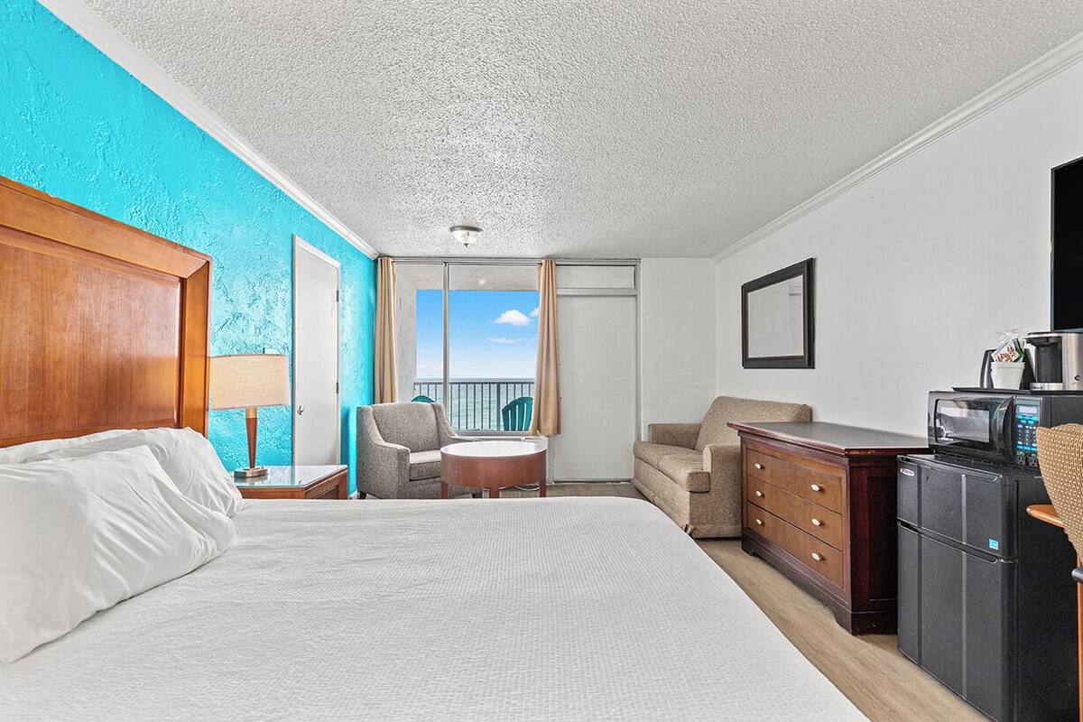 Ein Hotelzimmer mit einem großen Bett, einem Schreibtisch mit Kühlschrank, einem Sessel, einem kleinen Tisch und Blick auf den Himmel durch ein Fenster.