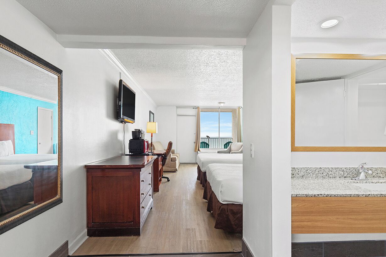 A hotel room with a wooden desk, a television, a bed, and a bathroom with a sink and mirror.