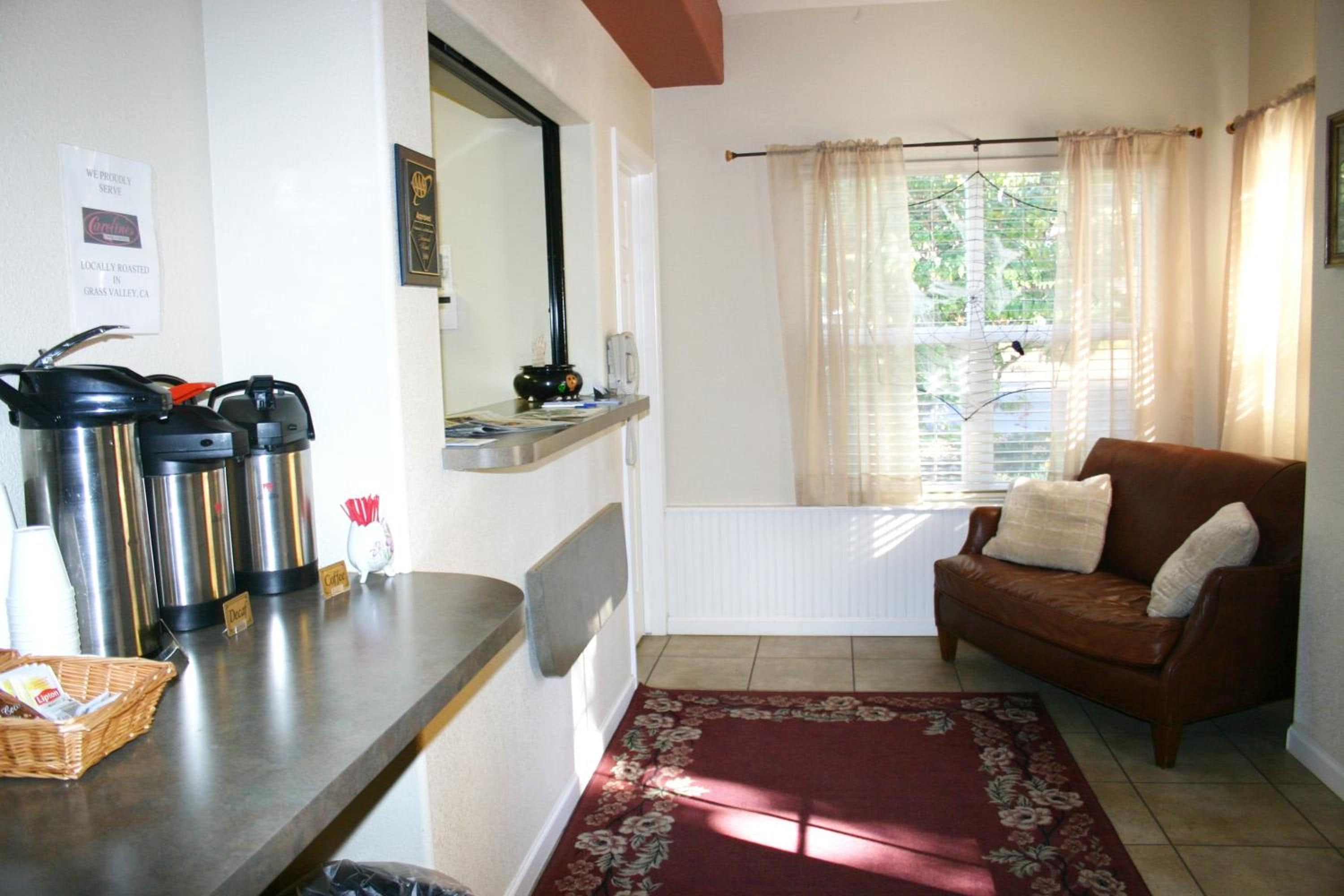 A cozy living room with a brown leather sofa, a coffee station with thermos cups, and a window with sheer curtains.