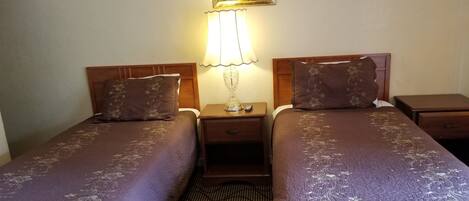 Ein Hotelzimmer mit zwei Betten, jedes mit einer blumigen Tagesdecke und passenden Kissen. Zwischen den Betten befindet sich ein Nachttisch mit einer Lampe, darüber an der Wand ein gerahmtes Pflanzenbild.