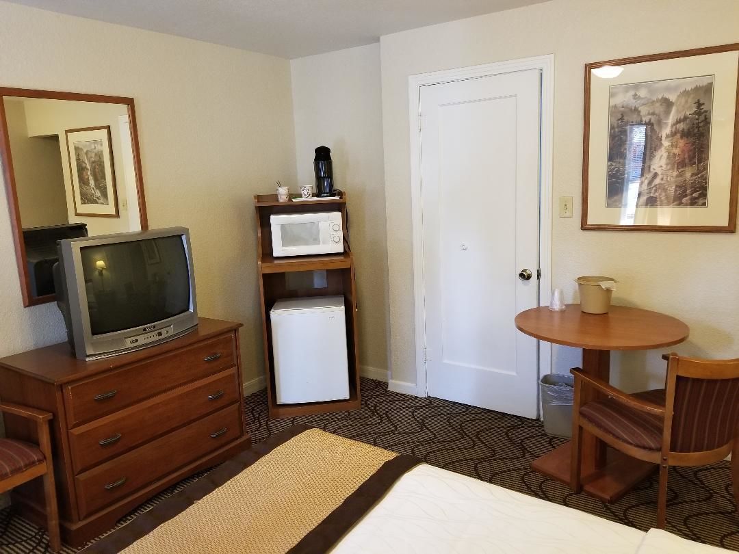 A hotel room with a wooden dresser, a television, a microwave, a small refrigerator, a dining table with a chair, and a door.
