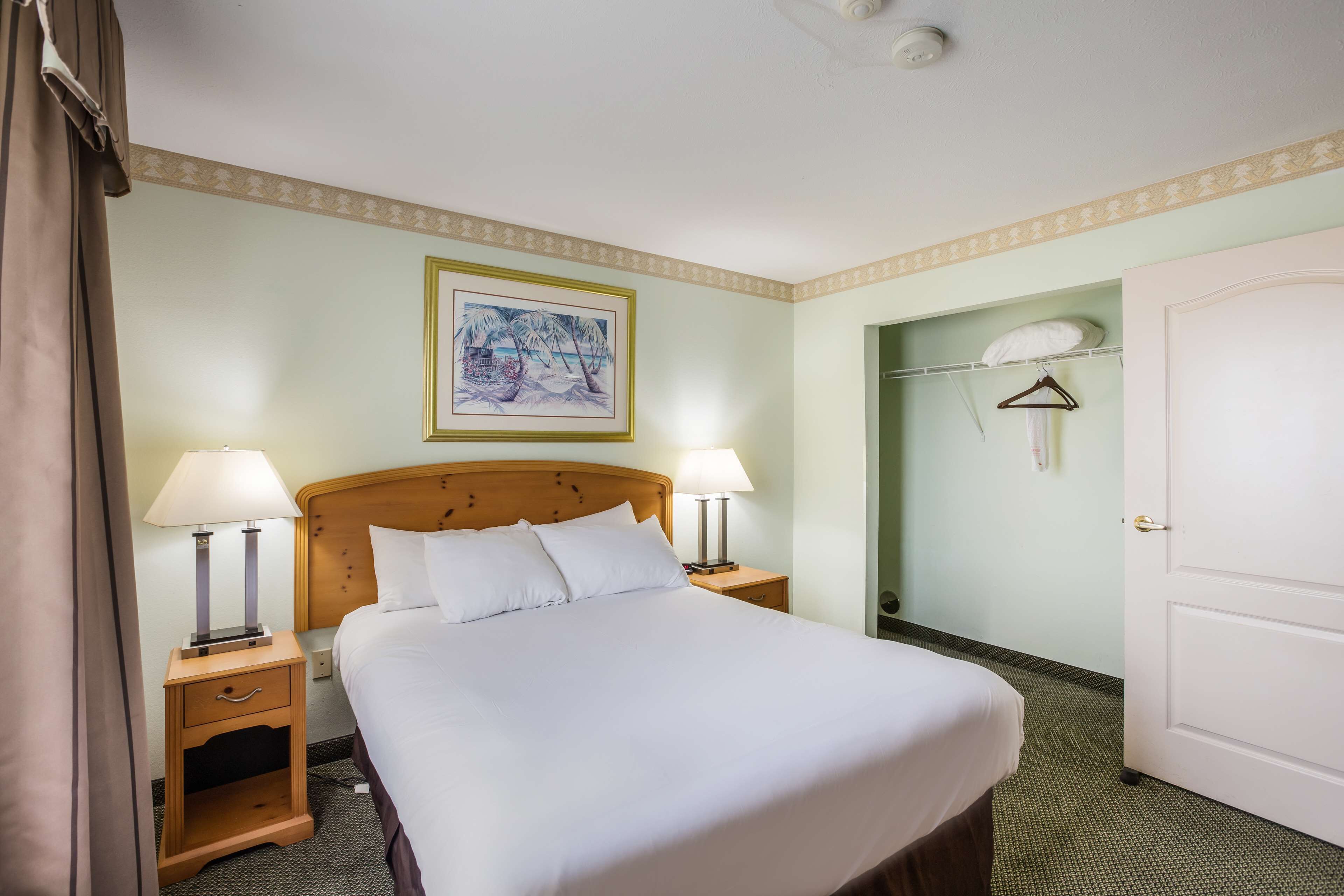 A neatly made bed with white linens, two bedside lamps, a wooden nightstand, a framed picture on the wall, and a door to a bathroom.